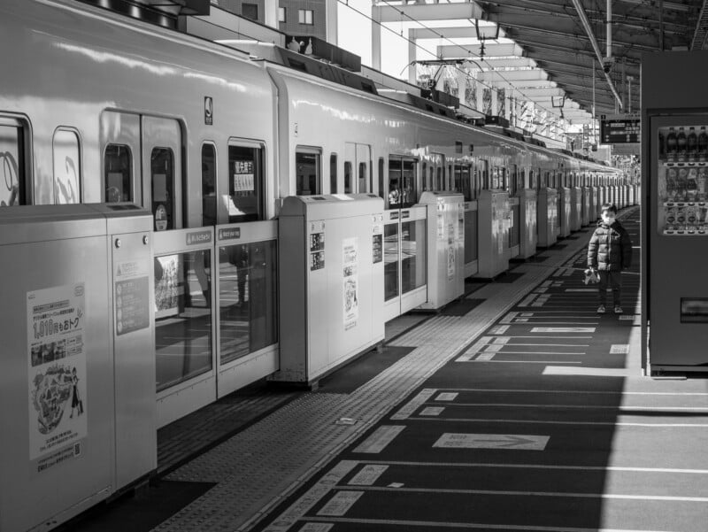 A black and white photo of a train station platform with a train stopped. A person wearing a mask stands alone in sunlight, while vending machines and signs line the platform.