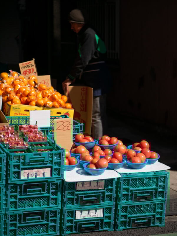 A street market stall displays neatly arranged crates of tomatoes and yellow fruits, with price signs in Japanese. A person in a beanie and jacket stands behind the stall, partially in shadow.