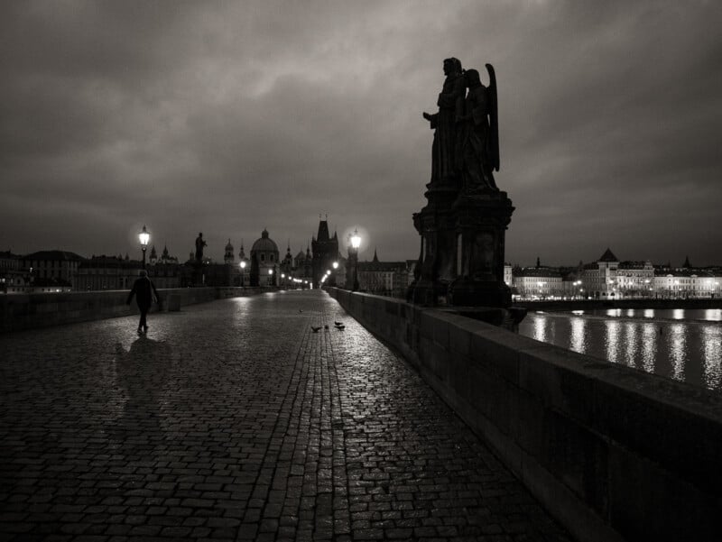 A person walks alone on a cobblestone bridge at dusk, passing large statues and street lamps. The city skyline and river are visible under a cloudy, moody sky.