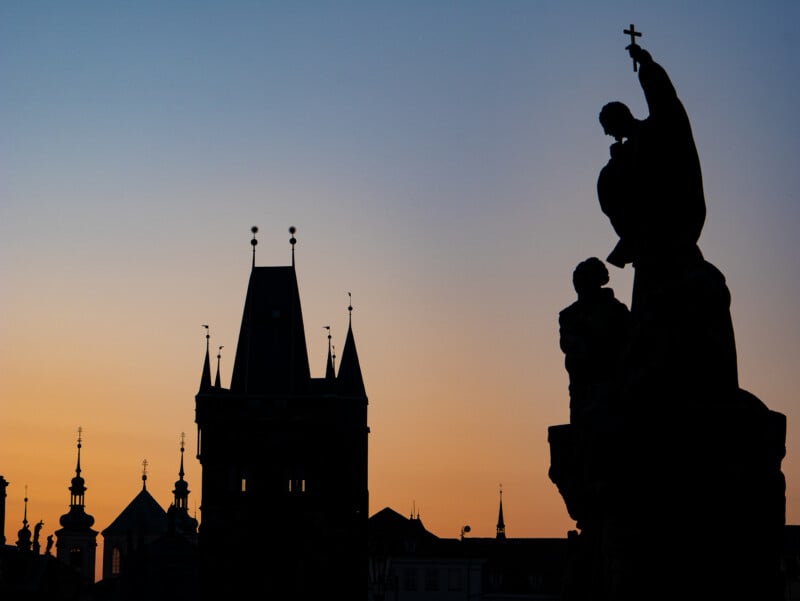 Silhouette of historic buildings and statues against an orange and blue sunset sky, with pointed towers and a figure holding a cross prominently in the foreground.
