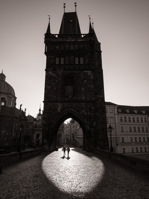 Two people walk hand in hand through an arched stone tower on a cobblestone bridge, illuminated by a bright circle of sunlight, with surrounding buildings in shadow.