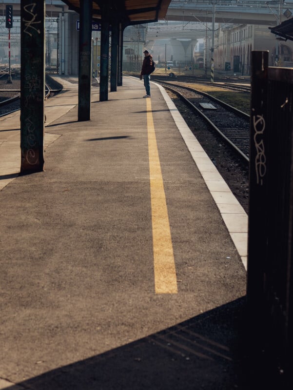 A person stands alone on a train station platform under a covered area, with sunlight casting long shadows. Train tracks curve alongside the empty platform, and graffiti is visible on nearby poles.