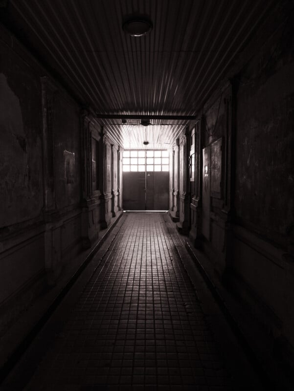 A dimly lit, narrow hallway with worn walls and a tiled floor leads to closed double doors with frosted windows, allowing some light to filter inside. The atmosphere is eerie and abandoned.
