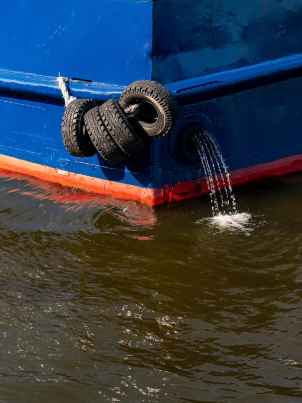 The bow of a blue and red boat with three tires hanging as bumpers. Water pours out from two circular outlets on the boat’s hull into the brownish water below.