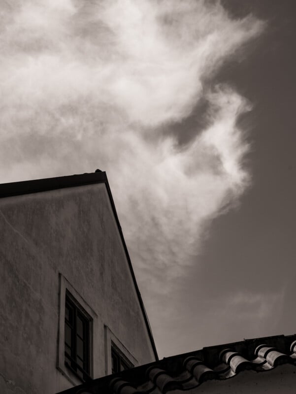 A sepia-toned photo shows the corner of a building with two windows and a tiled roof in the foreground, set against a sky with wispy, dramatic clouds.