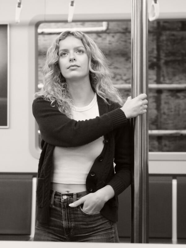 A woman with curly hair stands in a subway car, holding onto a pole with one hand and placing the other in her pocket. She wears a cardigan over a white shirt and looks thoughtfully ahead. The image is in black and white.
