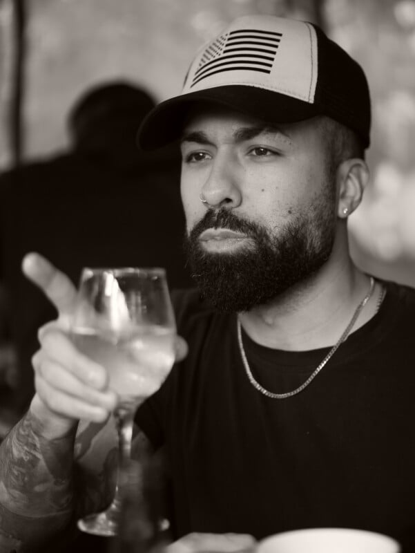 A bearded man wearing a cap and a chain necklace holds a glass of water and points with his finger. He appears to be deep in thought. The image is in black and white.