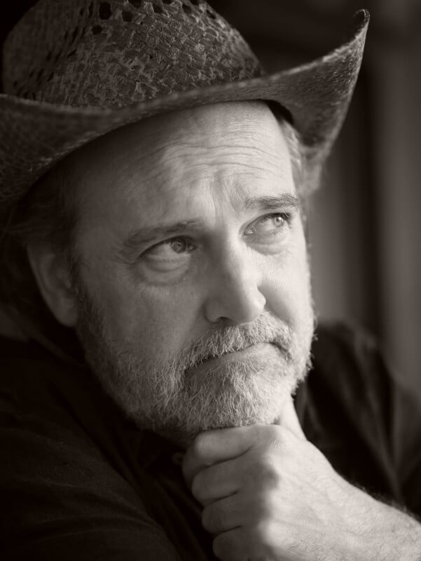 A middle-aged man with a beard and mustache wears a textured cowboy hat. He gazes thoughtfully to the side with his chin resting on his hand. The photo is in black and white.