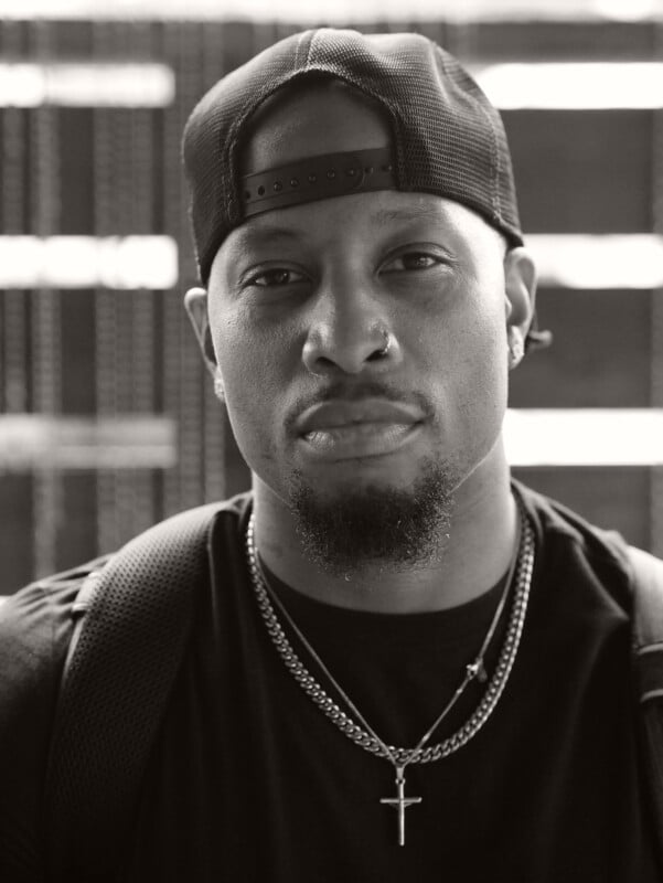 A man wearing a backward baseball cap, a nose ring, earrings, and layered necklaces, including a cross pendant, looks seriously at the camera. He has a goatee and a backpack. The photo is in black and white.