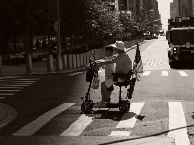 An older adult on a mobility scooter, carrying a U.S. flag, crosses a city street at a crosswalk with a younger companion. The scene is in black and white, with sunlight casting shadows on the road.