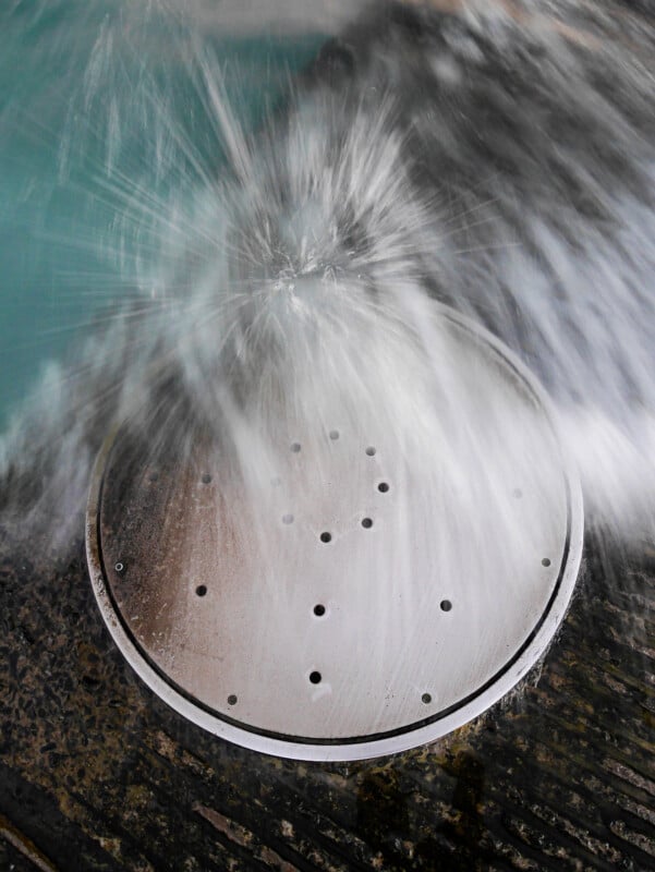 Water bursts upward and sprays out from a round metal drain cover with small holes, splashing across a wet, textured surface.