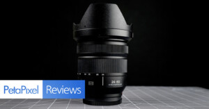 A wide-angle camera lens stands upright on a gray surface with grid lines, against a black background. "PetaPixel Reviews" is displayed in blue and white text at the bottom left.