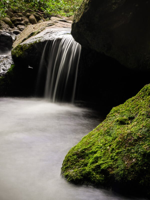 A small waterfall flows gently over a moss-covered rock into a calm, misty pool surrounded by lush greenery and smooth stones.