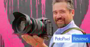 A smiling man with a beard holds a large Lumix camera towards the viewer, standing in front of a pink and black painted wall. The image features the PetaPixel Reviews logo in the lower right corner.