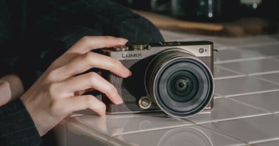 A hand rests on a white tiled surface, holding a Lumix digital camera with a large lens, positioned at an angle for a close-up view.