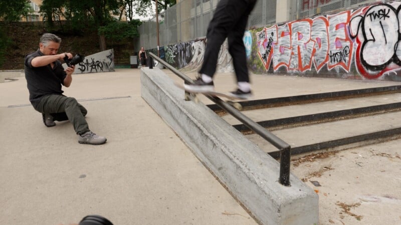 A photographer kneels at a skatepark, capturing a skateboarder grinding a rail. The background features graffiti-covered walls, trees, and another person standing near the fence.