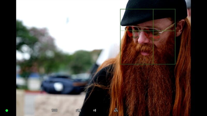 A person with long red hair and a thick beard wearing glasses and a black hat is photographed outdoors. The image has a focus rectangle overlay, indicating camera focus. Blurred cars and trees appear in the background.