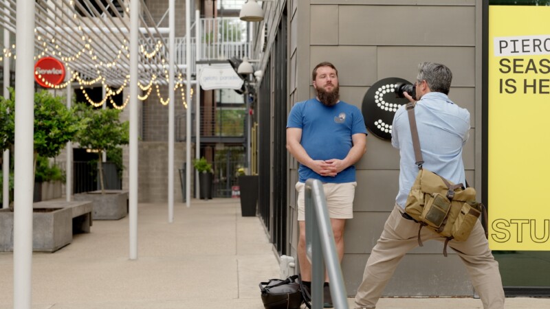A man in a blue shirt and shorts stands against a wall with a large letter "S" as another man with a messenger bag takes his photo on a quiet outdoor walkway.