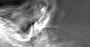 A black and white image of swirling gas and dust in space, with bright and dark areas creating a smoky, cloud-like effect, and small white stars scattered in the background.
