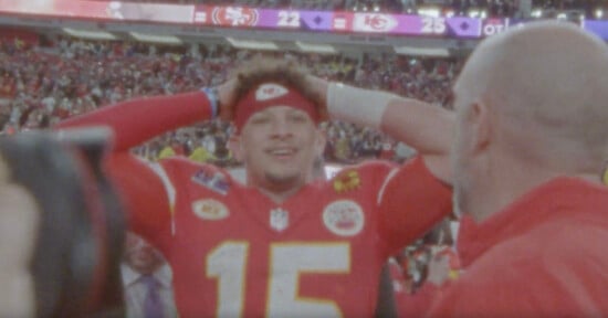 Patrick Mahomes wearing a red jersey with the number 15, and a red headband, looks joyful with his hands on his head amid a crowd in a stadium. The scoreboard in the background shows the 49ers 22 and Chiefs 25, suggesting a victory celebration.