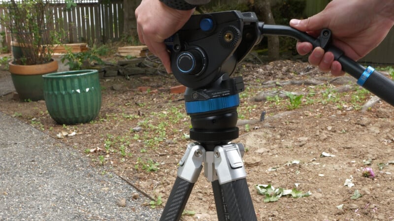 A person adjusts the pan handle of a professional video tripod outdoors on a dirt surface, with a green planter and wooden fence visible in the background.