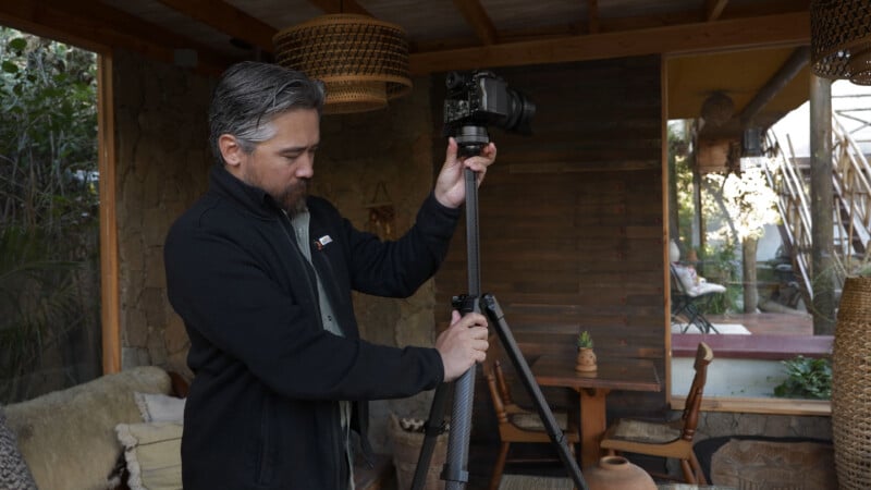 A man with gray hair and a beard adjusts a camera mounted on a tripod in a cozy, rustic indoor space with wood and stone decor.