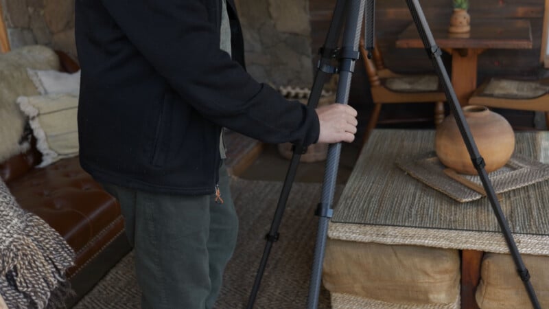 A person in a black jacket sets up a tripod beside a rustic table with a ceramic vase, surrounded by cozy furniture and wooden decor in a warmly lit room.
