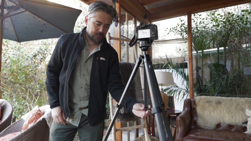 A man with gray hair and a beard adjusts a camera mounted on a tripod in a sunlit room with large windows, plants, and cozy furniture.