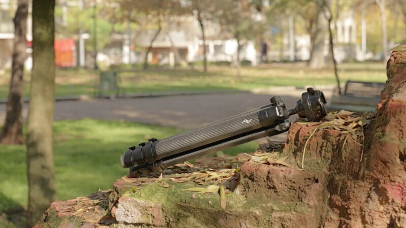 A black tripod is lying on a mossy brick ledge in a park with green grass, trees, and a blurred background of buildings and benches. Fallen leaves are scattered on the ledge and ground.
