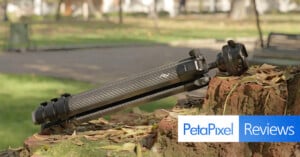 A black camera tripod rests on a mossy brick wall in a park, with fallen leaves around. The Petapixel Reviews logo is overlaid in the bottom right corner of the image.