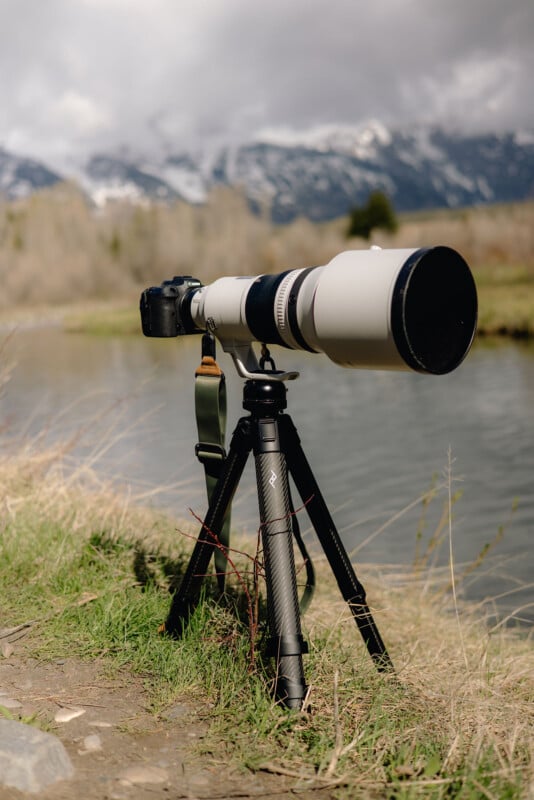 A large telephoto camera lens mounted on a tripod is set up by a river, with blurred mountains and trees in the background under a cloudy sky.