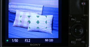 A camera screen displays a focused image of two patterned pillows on a blue-striped couch, with green focus brackets over the pillows and camera settings shown at the bottom.