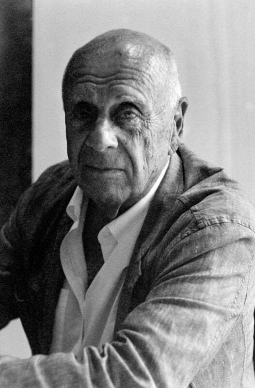 Black-and-white photo of an older man with short hair and a serious expression, wearing a light-colored shirt and a textured jacket, sitting and looking directly at the camera.