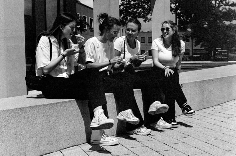 Four young women sit on a stone bench outdoors, smiling and talking while eating ice cream. They are casually dressed, wearing light tops and dark pants, and appear relaxed and happy in the sun.