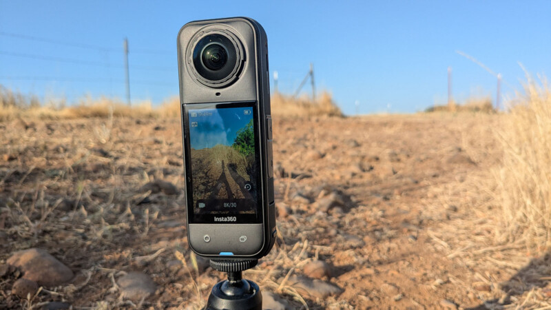 A 360-degree action camera mounted on a tripod stands on a dry, rocky dirt path with tall, yellow grass on both sides and a clear blue sky in the background.