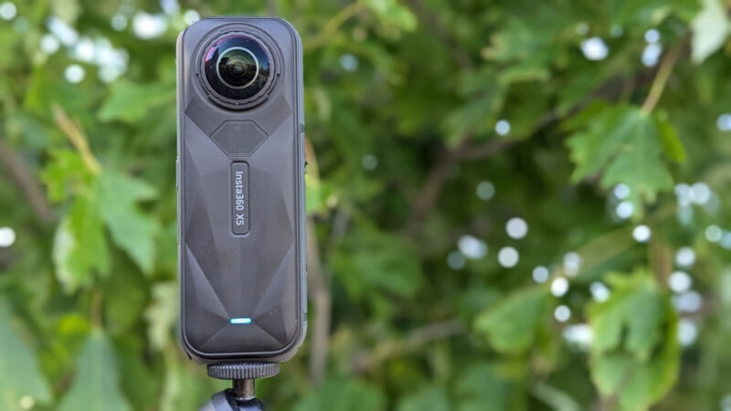 A black 360-degree camera mounted on a tripod stands in front of green leafy foliage. The camera lens is prominent at the top. The background is blurred.