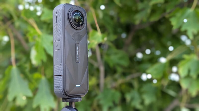 A black Insta360 X3 action camera mounted on a tripod stands outdoors in front of leafy green foliage.