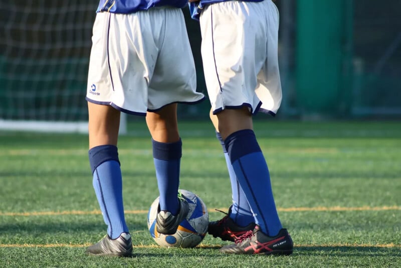 Two soccer players in blue socks and white shorts stand on a grassy field, with one balancing a soccer ball between their feet. A goal net is visible in the background.