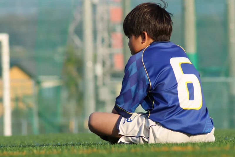 A young boy in a blue soccer jersey with the number 6 sits on grass, facing away, with his knees up and arms resting on them, on a sunny day at a sports field.