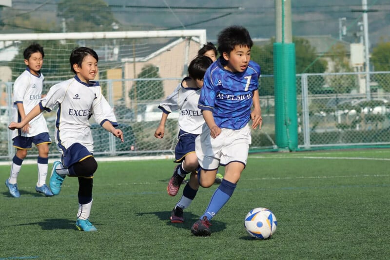 A group of young boys play soccer on a field. A boy in a blue jersey runs ahead, dribbling the ball, while several boys in white jerseys chase after him. The scene is energetic and takes place outdoors.
