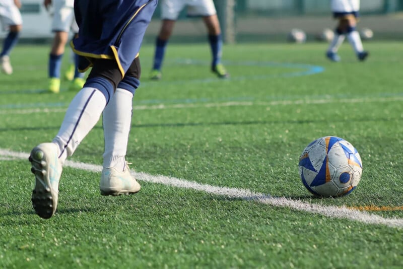 A soccer player in white socks and blue shorts runs toward a blue and white soccer ball on a green artificial turf field, with other players visible in the background.