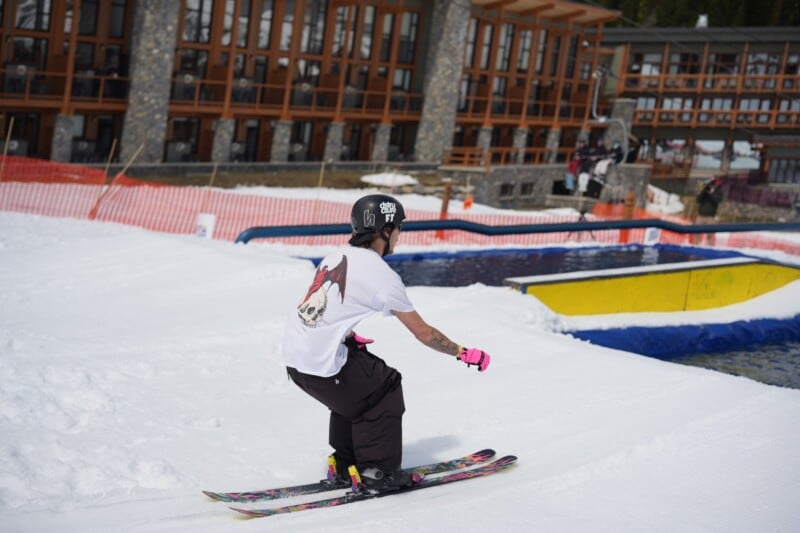 A skier wearing a helmet, white t-shirt, black pants, and pink gloves prepares to jump over a pool of water on a snowy slope near a lodge with large windows.