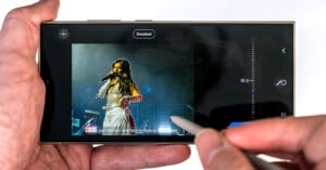 A hand holding a smartphone with a stylus, editing an image of a female singer on stage, highlighting her figure with a selection tool on the screen.
