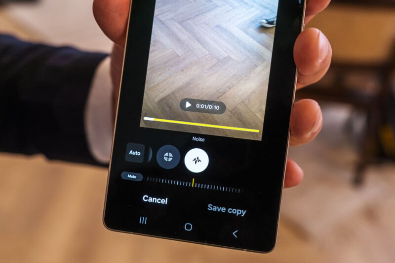 Close-up of a person holding a smartphone, displaying an audio editing app. The screen shows a noise reduction feature with a yellow progress bar. The background includes a wooden herringbone-patterned floor.