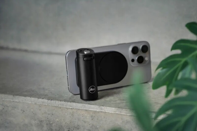 A smartphone with a triple camera and a circular accessory attached is placed on a gray surface. In front of it is a small black cylindrical device, and a green plant leaf is partially visible on the right.