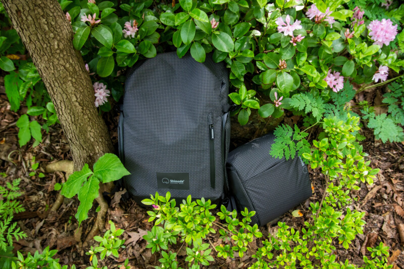 A black textured backpack and a matching smaller pouch rest on the ground amid green foliage, flowering bushes, and tree trunks in an outdoor garden setting.