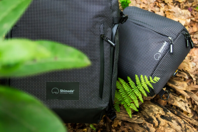 Two Shimoda-branded black backpacks with a grid pattern rest on the forest floor among green leaves, a fern, and dried brown foliage.