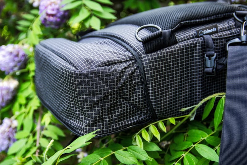 A close-up of a black, grid-patterned Shimoda camera bag resting among green leaves and purple flowers, with its strap and zippers visible.