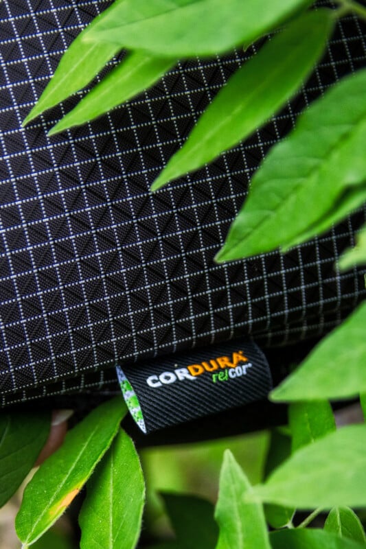 A close-up of a black Cordura fabric bag with a white grid pattern and a visible "CORDURA re/cor" tag, partially covered by green leaves.