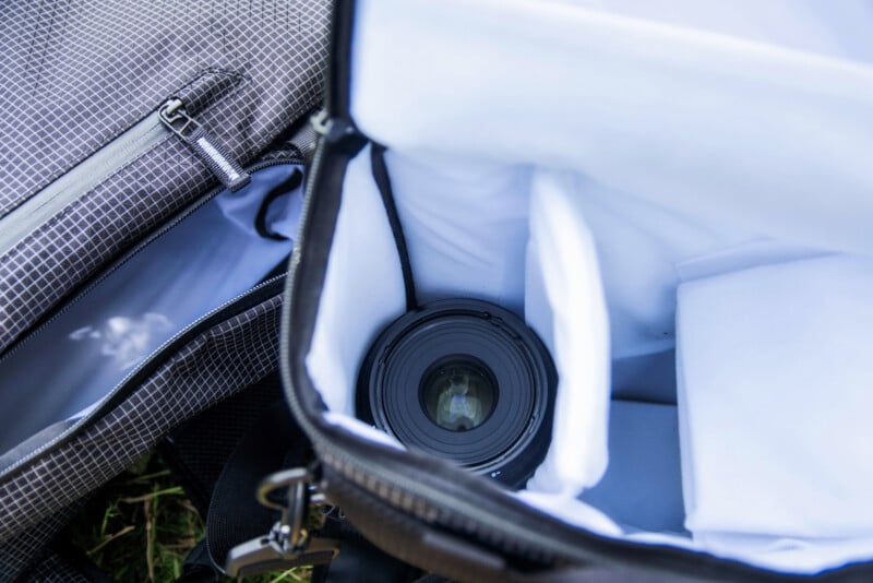 Open camera bag with padded dividers, showing a camera lens stored inside. The bag’s zippered pocket and soft white interior lining are also visible.